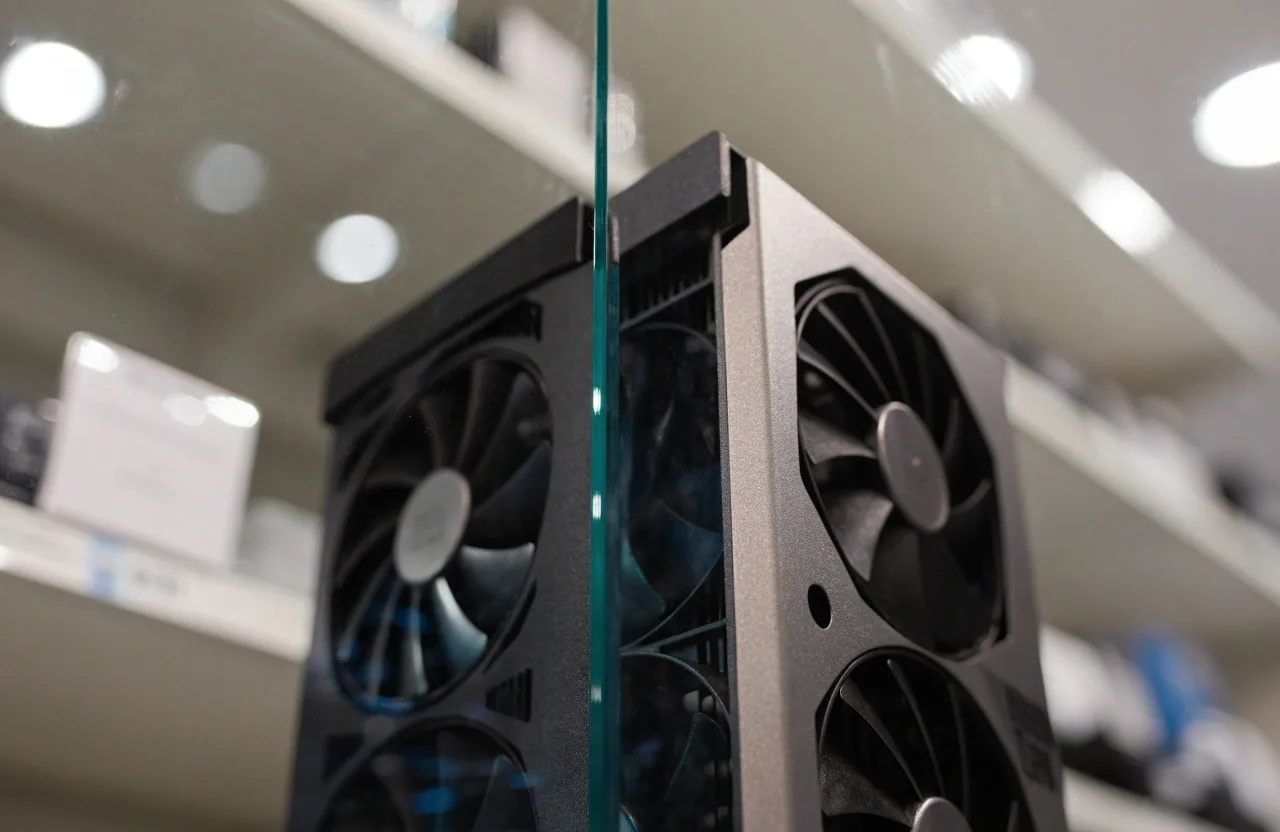 Close three-quarter view of a glossy acrylic display panel face in a bright electronics hall with a graphics card box standing upright behind it, printed surface turned away from the camera