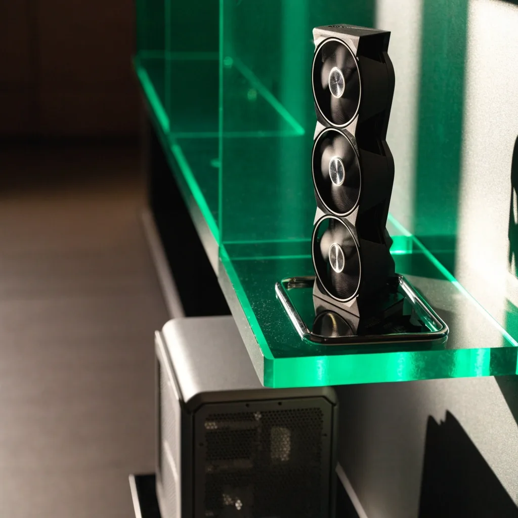 An upper-midrange triple-fan graphics card on a chrome tray inside a lit acrylic shelf, with a small PC case beside it for size reference